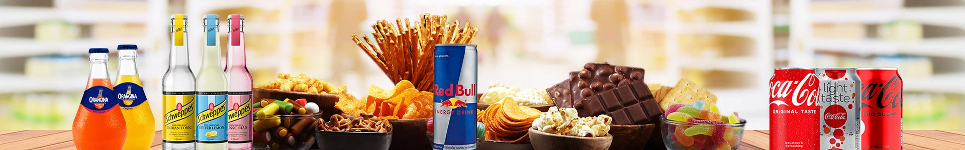 Getränke und Snacks im Sortiment: Orangina, Schweppes, Red Bull, Coca-Cola sowie Chips, Salzstangen, Schokolade, Popcorn und Gummibärchen auf einem Holztisch mit unscharfem Hintergrund.