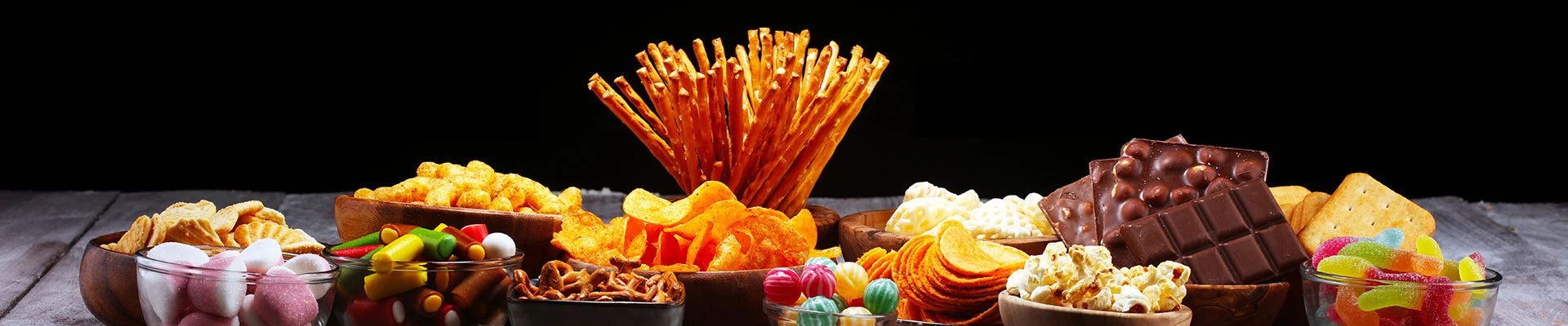 Vielfältige Snackauswahl mit Chips, Salzstangen, Popcorn, Schokolade, Keksen, Gummibärchen und bunten Süßigkeiten in Schalen auf einem Holztisch.